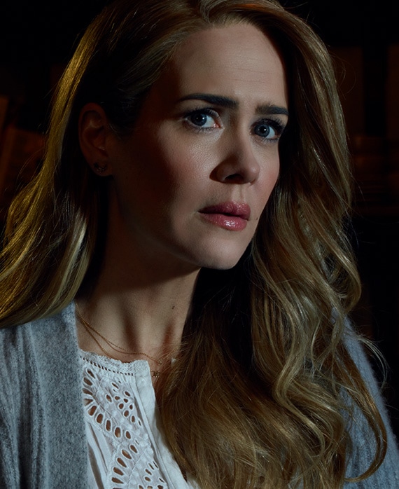 Sarah Paulson headshot in white lace patterned top and grey cardigan with hair down over shoulder and light illuminating face