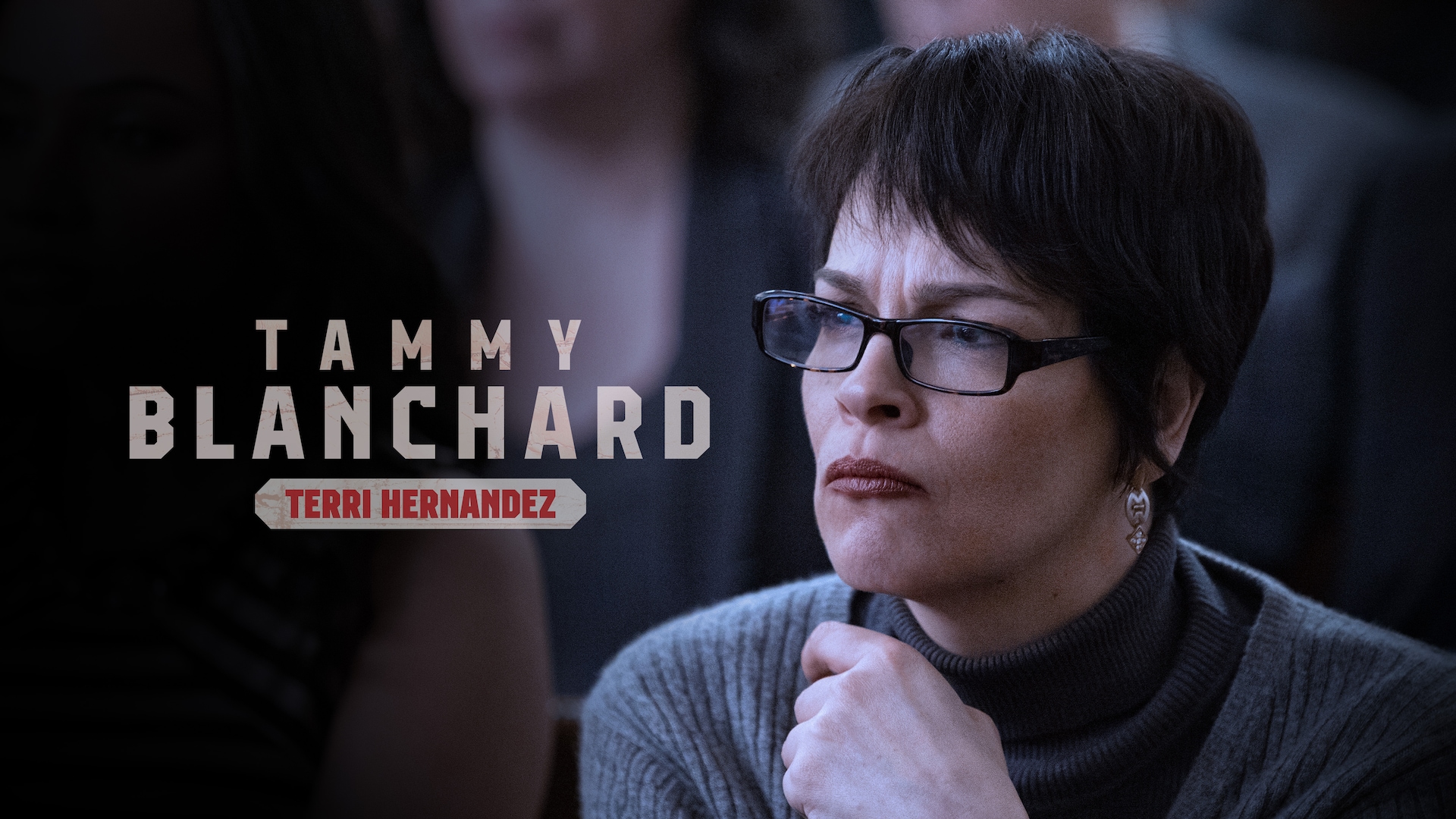 Tammy Blanchard as Terri Hernandez in a grey sweater and black turtleneck shirt, wearing thin framed glasses, sitting.