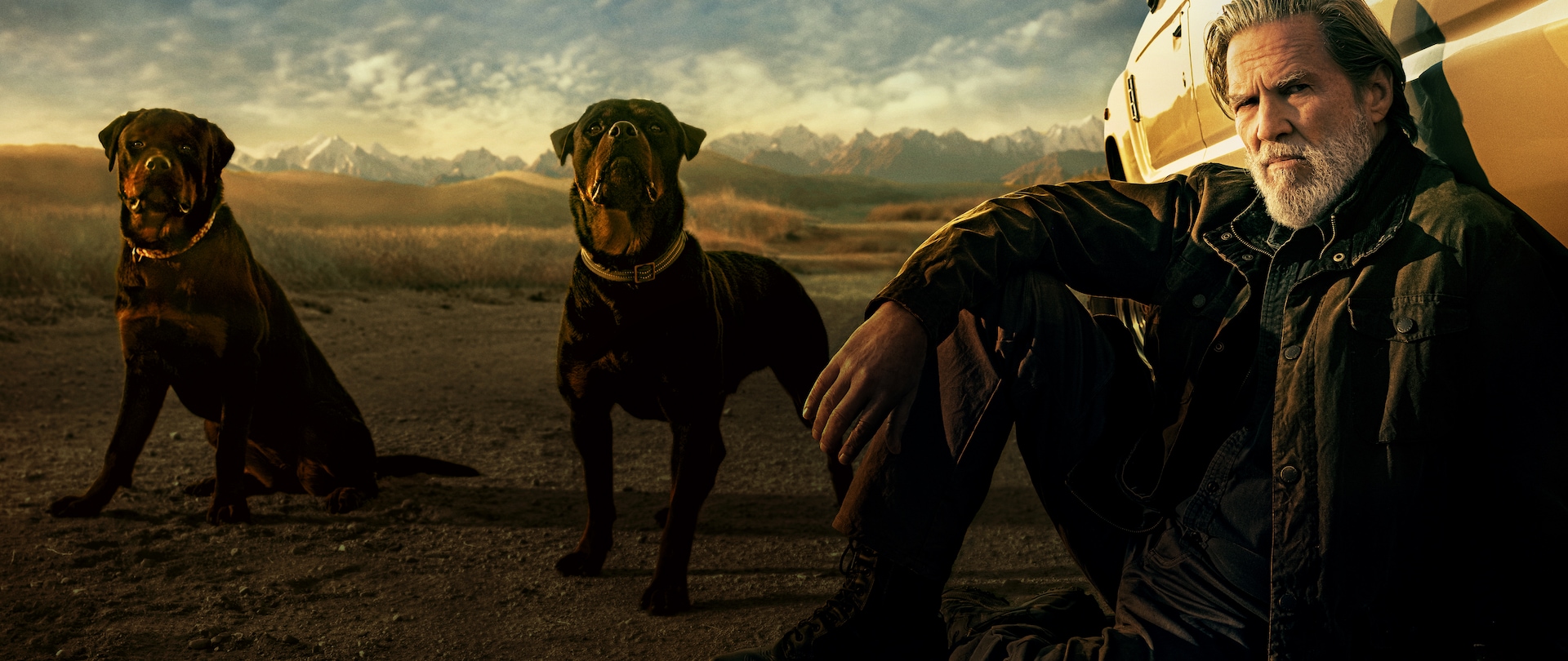 Jeff Bridges as Dan Chase sitting on a gravel road in middle of a desert, leaning on a car with two Rottweilers next to him