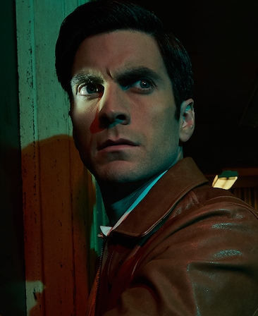 Wes Bentley headshot in brown leather zipper jacket outside by wooden post at night with right eye illuminated