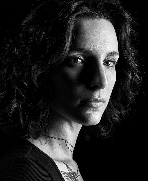 Carla Baratta headshot with tattoos on neck and chest and wearing a black shirt