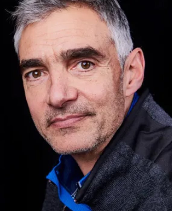 Headshot of Peter Ligouri in a grey coat and a bright blue top poking out, in front of a black background.