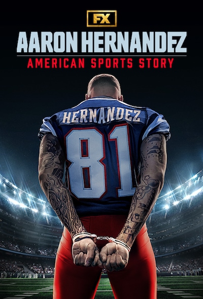 Aaron Hernandez in blue and white football uniform with hands behind his back in handcuffs standing in a stadium at night.