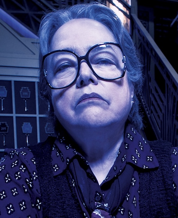 Kathy Bates headshot in purple patterned button down and big wide black glasses in front of staircase under purple lighting
