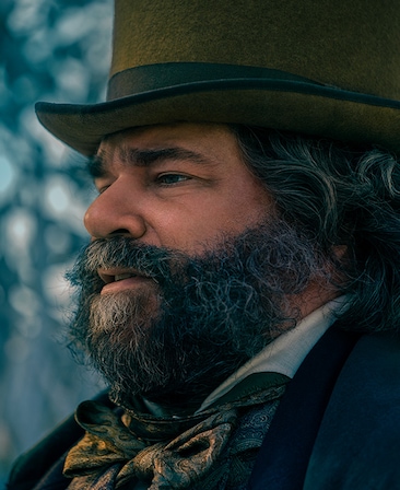 Matt Berry headshot of side profile in winter coat with ornate bowtie neck scarf with olive green top hat