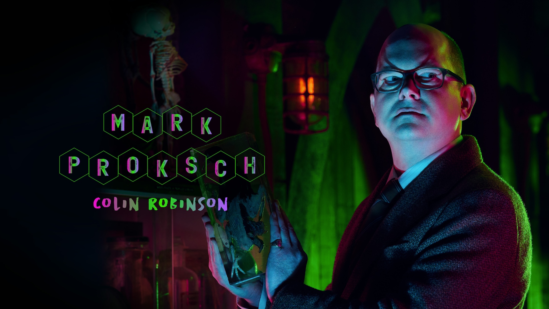 Mark Proksch as Colin Robinson holding a jarred specimen in a pink and green lit laboratory.