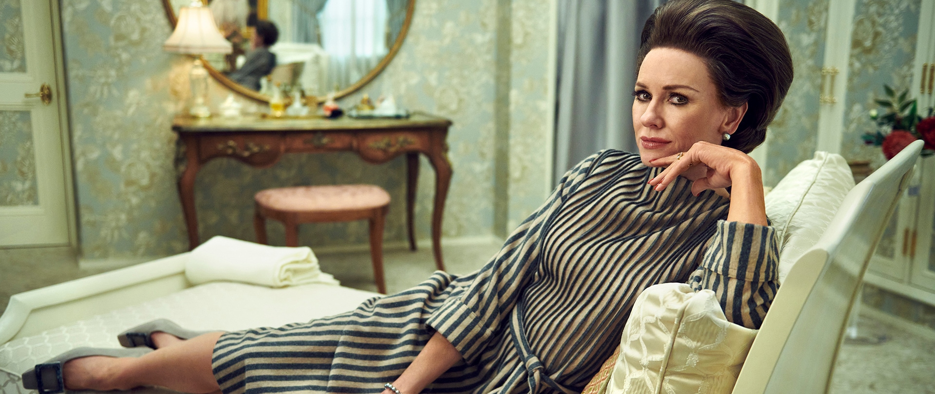 Naomi Watts as Babe Paley in Feud wearing a striped dress laying on a chaise in an elegantly styled home.