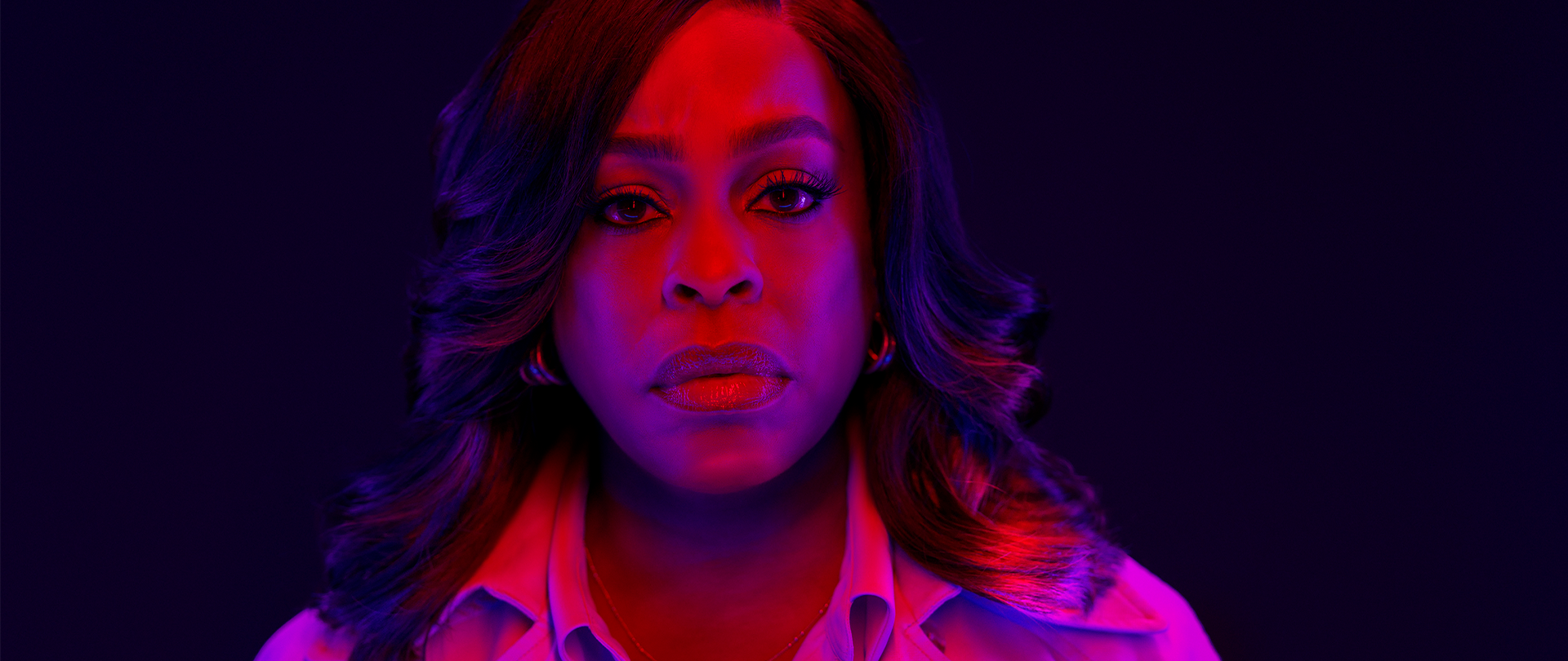 Niecy Nash-Betts as Lois Tryon in FX's Grotesquerie in a button up and coat with pink and purple lights shone on her face.