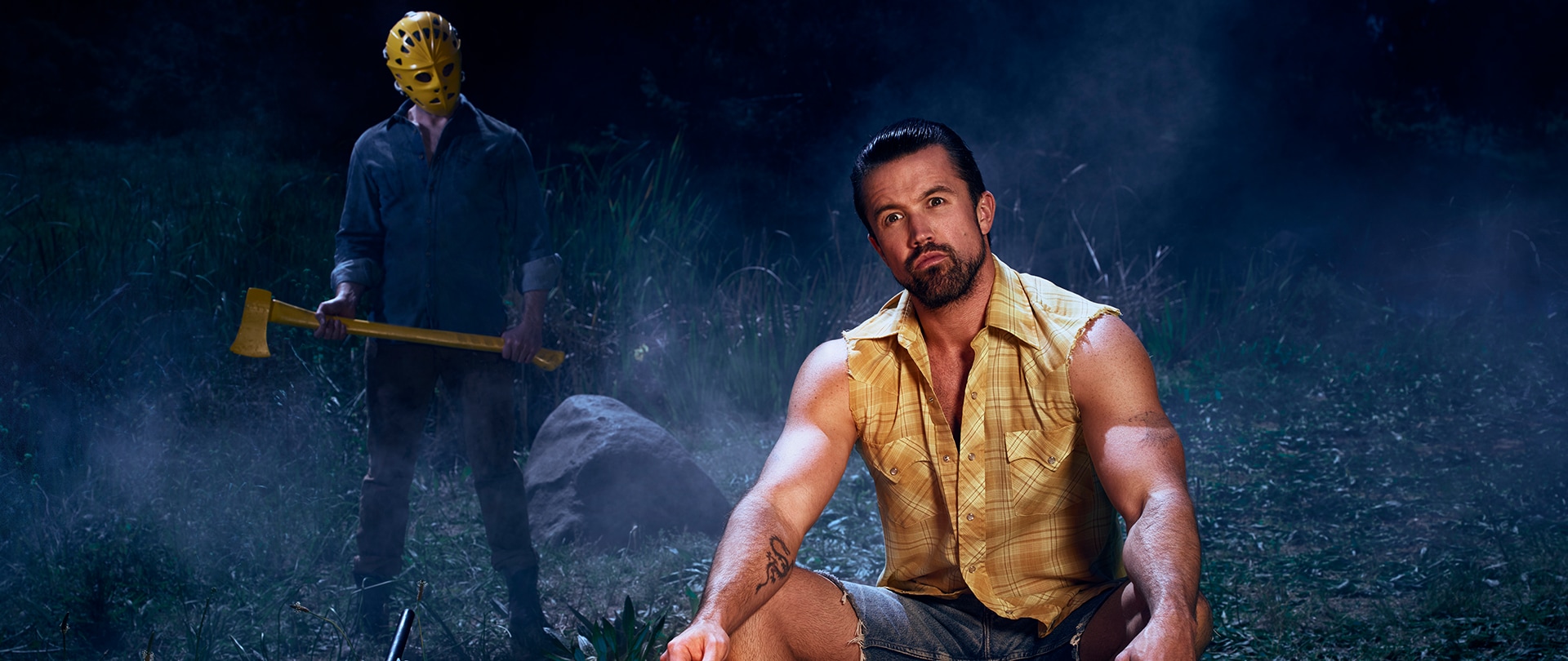 Rob McElhenney as Mac in a ripped plaid shirt at a campfire with a person in a mask holding an axe behind him.