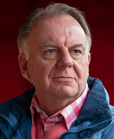 Paul Clayton wearing a pink and white plaid shirt with a blue jacket standing in front of a red background