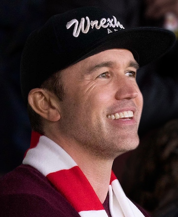 Rob McElhenney headshot wearing a red and white scarf and black Wrexham hat