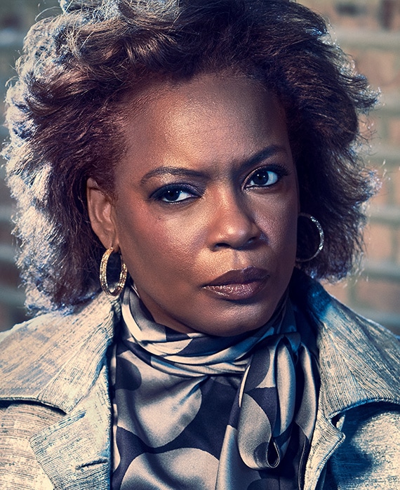 Aunjanue Elis headshot wearing a patterned shirt and earrings standing in front of a brick wall