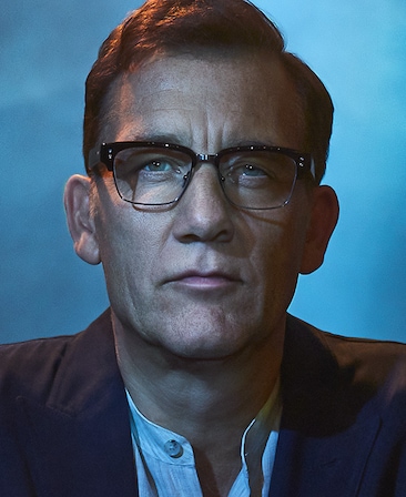 Clive Owen wearing glasses and a suit standing against a blue background