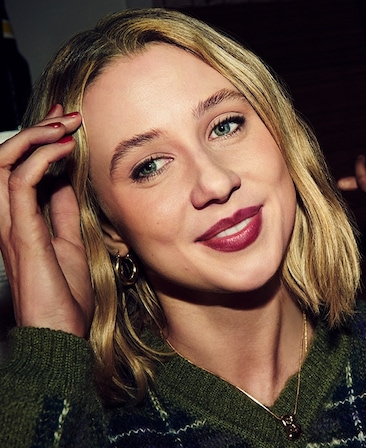 Headshot of Lucy Freyer as Billie in FX's Adults wearing a green plaid sweater and gold necklace, with her head angled. 