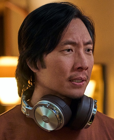 Headshot of Kelvin Yu playing Noah with head phones around his neck in a brown shirt and yellow background.