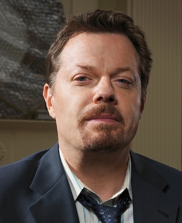 Eddie Izzard headshot wearing a navy tie, striped shirt and black jacket, standing in front of a picture