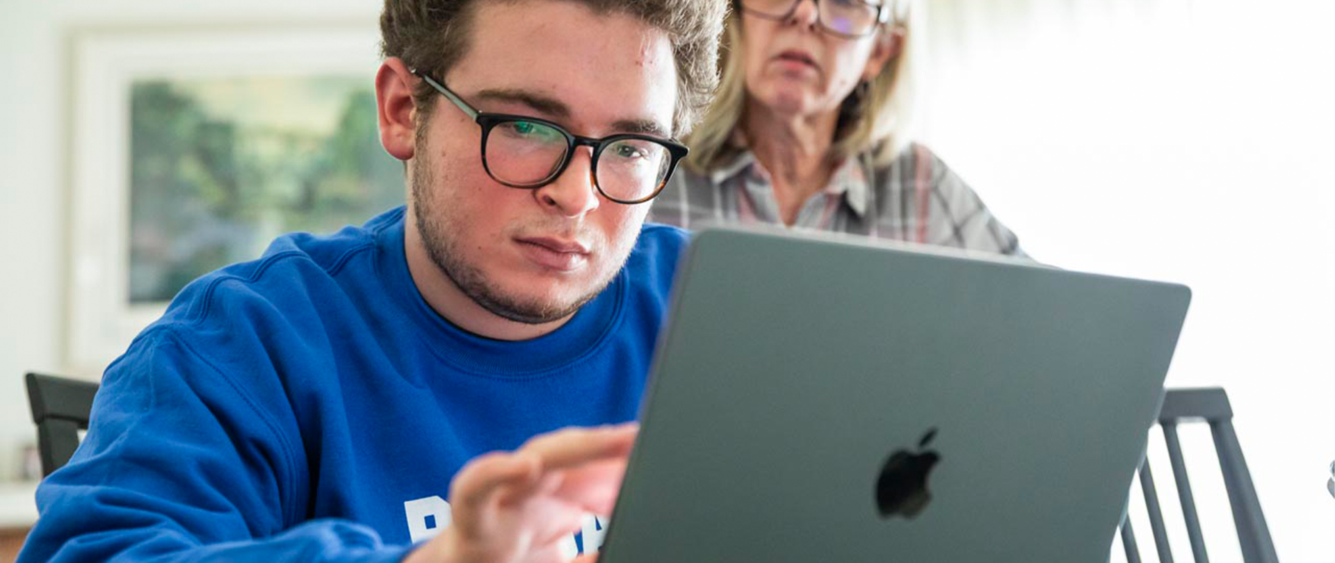 Boy with glasses and a blue sweatshirt typing on a laptop.