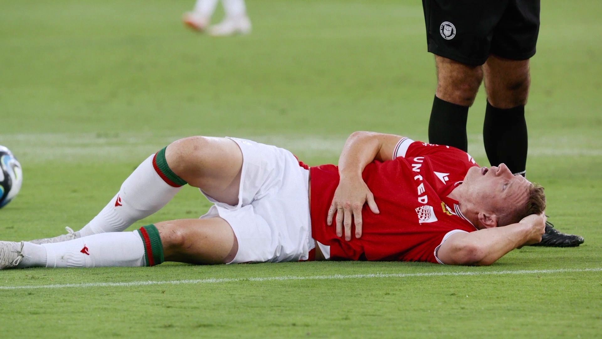 A football player wearing a red and white jersey laying on the football field with a soccer ball behind him.
