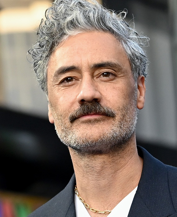 Taika Waititi headshot wearing a white shirt with black jacket and chain necklace.