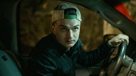 Man wearing a backwards hat sitting in the driver's seat of a dimly lit car. 