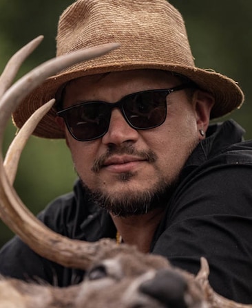 Sterlin Harjo headshot wearing black glasses and a woven tan hat, with a deer in front of him.