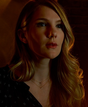 Lily Rabe headshot with long blonde hair parted over her left shoulder in black v neck dress and dainty gold necklaces