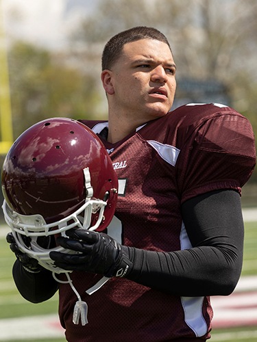 Hernandez wearing a burgundy football uniform and holding a helmet looks to the side of the field
