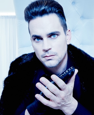 Matt Bomer with hair styled up and wearing black fur jacket with black latex glove over one hand while clasping hands