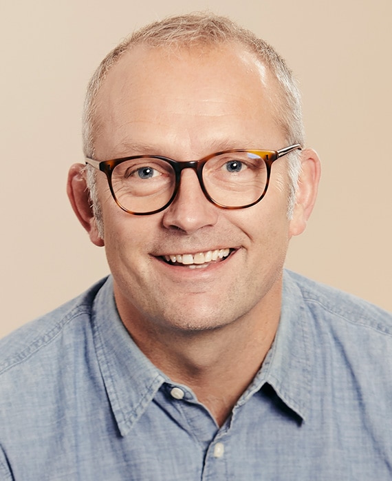 Rob Aslett headshot in light blue denim button down with light peach background