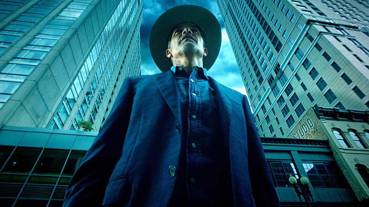 Raylan Givens wearing a suit and denim shirt and standing in front of two skyscrapers