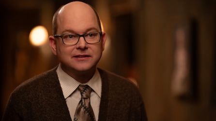 Mark Proksch as Colin Robinson in a brown suit in a dimly lit room.