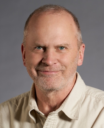 Sam Johnson headshot wearing a beige button down shirt, standing in front of a gray background.