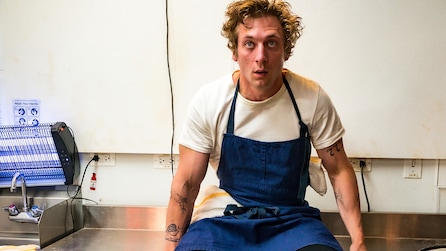 Jeremy Allen White as Carmy wearing blue apron sitting on restaurant kitchen table in FX's The Bear 
