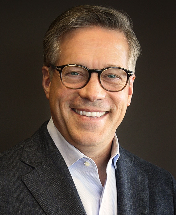Steve Stark headshot wearing a white button down and black suit jacket with glasses, against a black background