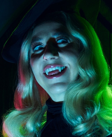 Headshot of Kristen Schaal as The Guide wearing a black hat and fangs as teeth.