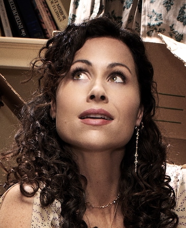 Minnie Driver headshot wearing a long earring and necklace with books on a shelf behind her