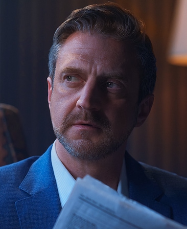 Raul Esparza headshot wearing a suit and holding a newspaper
