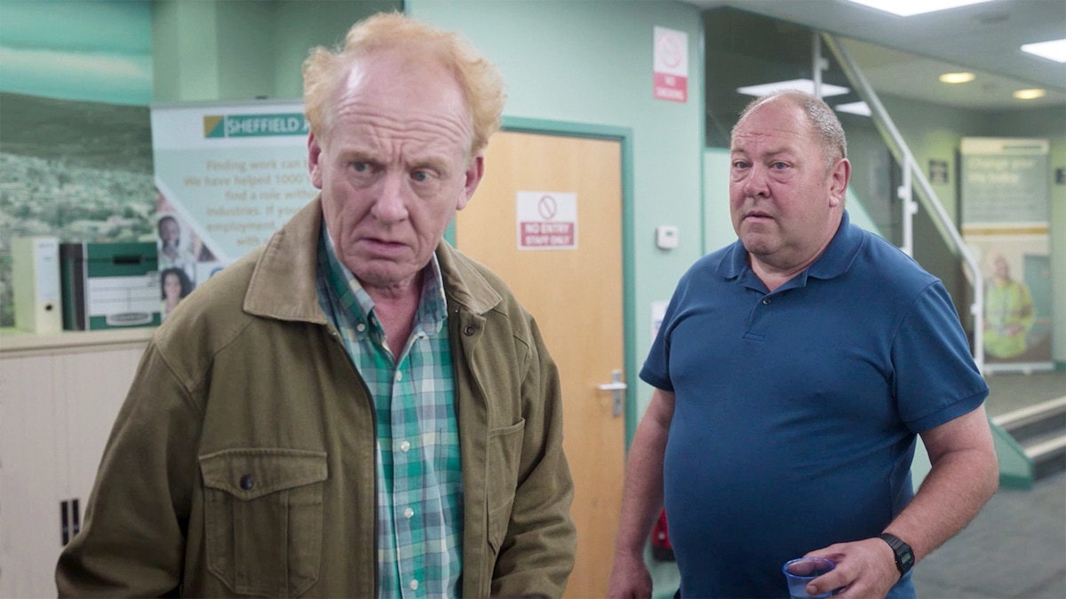 Two men inside a building,, on wearing a green paid shirt and the other a blue polo shirt