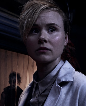 Alison Pill in white button down and coat in dark room looking up with clown with messy hair behind her by plastic covers