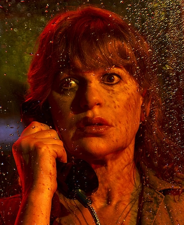 Headshot of Sandra Bernhard talking on payphone by rainy window and red lighting