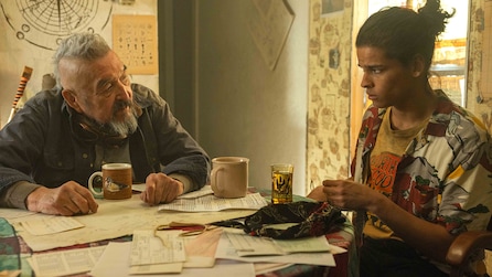 Bear, sitting at a table covered in papers, with another man drinking coffee
