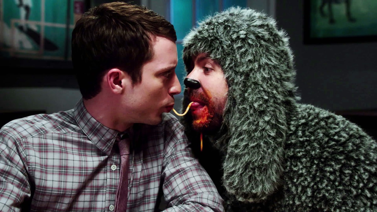 Man in dog costume sharing a noodle with another man with their mouths