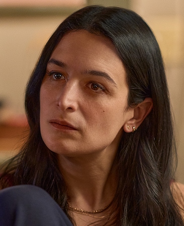 Headshot of Jenny Slate as Nikki Boyer sitting with her knee to her chest, wearing gold jewelry.