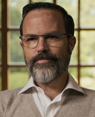 Michael Williamson headshot wearing a brown sweater over a white button up, in beige rimmed glasses, in front of a window.
