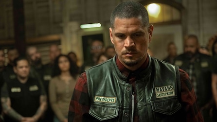 EZ wearing a leather vest that reads "presidente" while a group of people stand behind him.