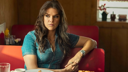 Rita sits in a diner wearing a blue denim shirt. 
