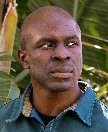 Gbenga Akinnagbe headshot wearing a blue collar shirt with foliage behind him.