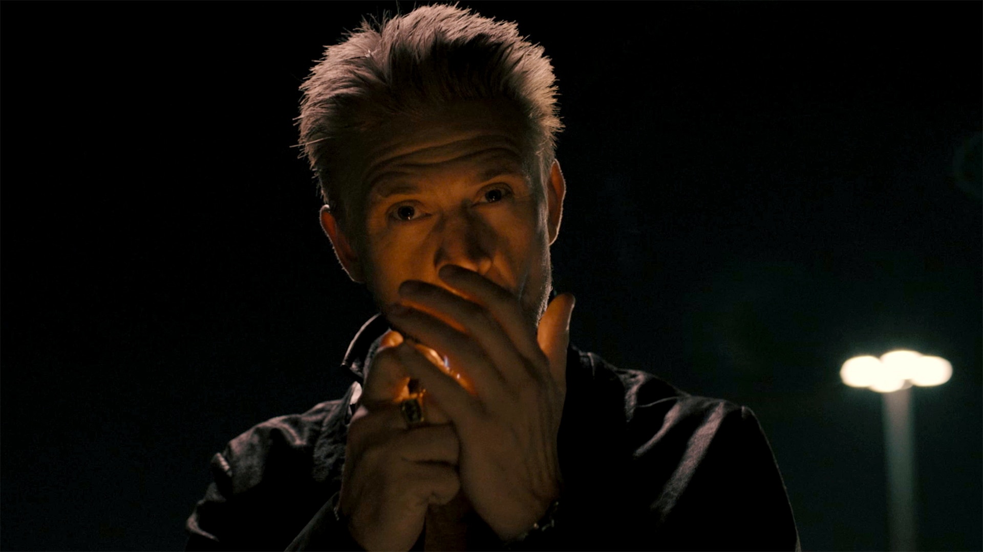 Raylan Givens lighting a flame behind his hands while standing in the dark