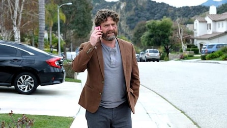 Zach Galifianakis in brown blazer talking on red phone on residential sidewalk in FX's Baskets
