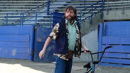 Zach Galifianakis standing on floor of empty stadium holding a bike during the day in FX's Baskets